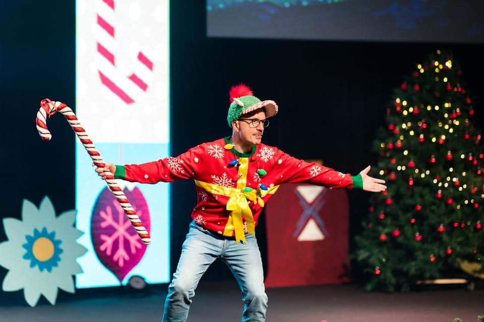 A guy on stage decked out in Christmas gear holding a candy cane. This images is used for our header image for our blog, "10 Engaging Christmas Games for Kids Ministry This Season."