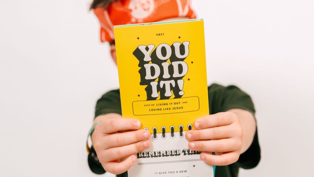 Blog is of a kid with a Vacation Bible School journal saying "you did it!" This image is being used for the blog header for the blog, Your Ultimate Guide to Marketing a Memorable VBS